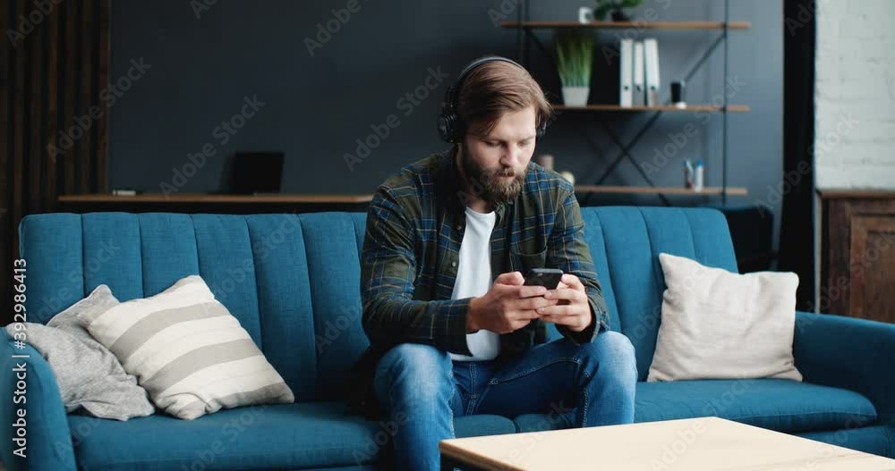Young attractive Male is enjoying the Music, wearing Headphones, and texting someone through an App on his Phone. Portrait of a young man spending time, having Fun at Home. Apps. Music. Indoor. Young attractive Male is enjoying the Music, wearing Headphones, and texting someone through an App on his Phone. Portrait of a young man spending time, having Fun at Home. Apps. Music. Indoor.