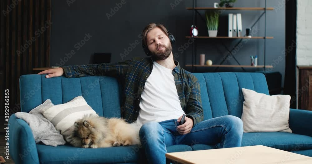 Calm Relaxed Caucasian Bearded Man is enjoying while listening to his favorite Music. Young Male is spending Time at Home, Playing with his Cute Dog on the Couch. Wearing Headphones. Music Apps. Calm Relaxed Caucasian Bearded Man is enjoying while listening to his favorite Music. Young Male is spending Time at Home, Playing with his Cute Dog on the Couch. Wearing Headphones. Music Apps.