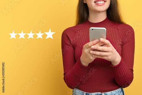 Woman leaving review online via smartphone on yellow background with five stars, closeup. Service or product feedback photo