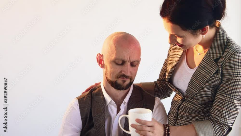 Family business. Beloved couple. Break time. Interest support. Stylish man working laptop taking cup coffee with pleasure bringing by wife hugging him back white background slide shot. Family business. Beloved couple. Break time. Interest support. Stylish man working laptop taking cup coffee with pleasure bringing by wife hugging him back white background slide shot.