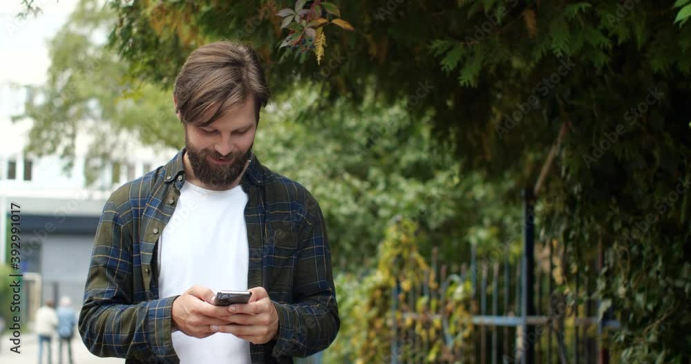 Portrait of Cute Caucasian Young Man is walking in Park, Texting Messages. Handsome Hipster Guy is walking outside on the Warm Day, browsing his Cell Phone, having Fun. Good Mood. Apps. Outdoor. Portrait of Cute Caucasian Young Man is walking in Park, Texting Messages. Handsome Hipster Guy is walking outside on the Warm Day, browsing his Cell Phone, having Fun. Good Mood. Apps. Outdoor.