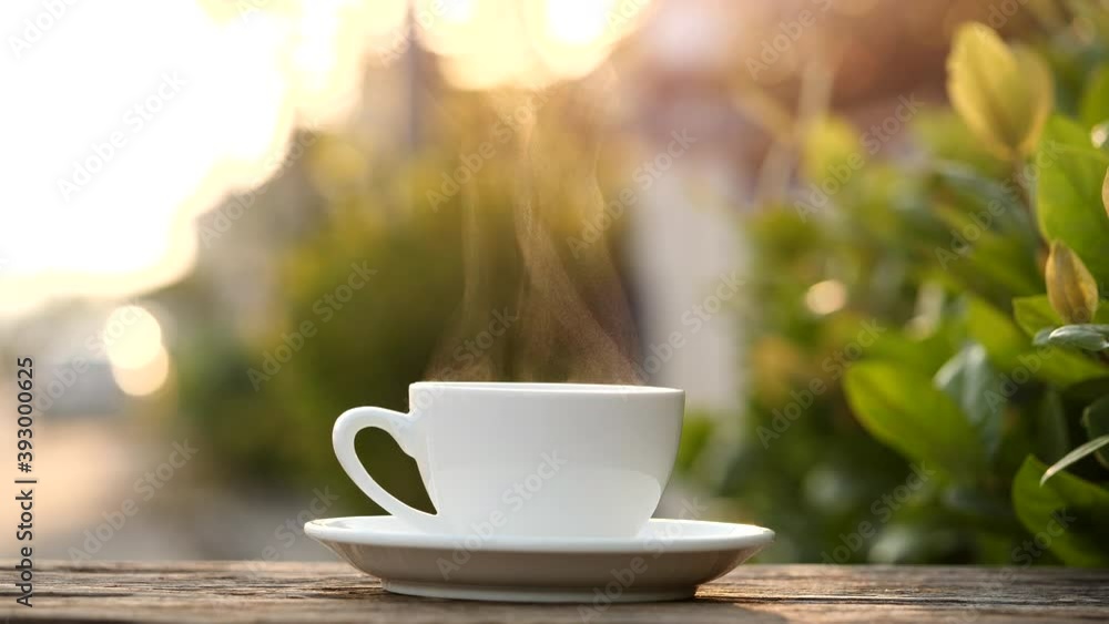Close-up steaming hot coffee or tea cup on saucer, slow motion. Hot ceramic white coffee cup with smoke on old wooden table in nature background. Hot Coffee Drink Concept