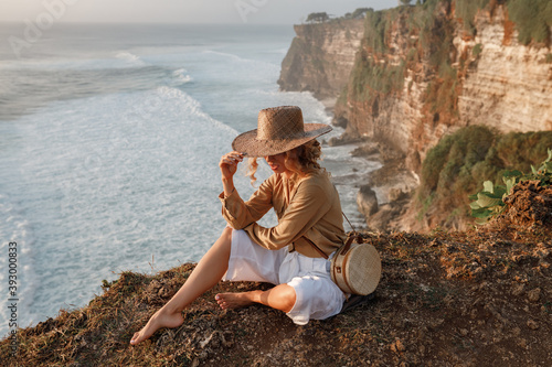 Young tourist woman in straw hat, cover the face, sit on the edge of a cliff with sea view. Travel Bali, explore beautiful places of the island