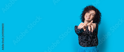 Curly haired woman is pointing at camera and smile on a blue studio wall with free space