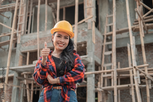 beautiful female contractor with thumbs up wearing a safety helmet in the background of building construction