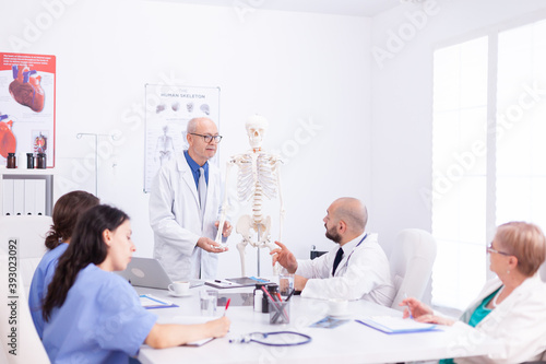 Mature doctor wearing glasses while holding presentation about human anatomy using skeleton. Clinic expert therapist talking with colleagues about disease, medicine professional