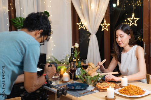 Young Asian man in blue t-shirt and eyeglasses recording video of young Asian beautiful girl in white sleeveless dress preparing food on dining table for her personal vlog, cooking and dinner vlog.