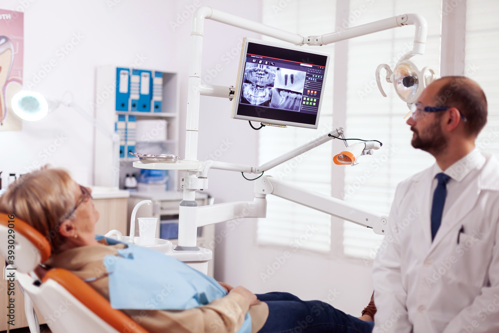 Obraz premium Stomatolog talking about teeh treatment to senior woman in dentist cabient. Medical teeth care taker pointing at patient radiography on screen sitting on chair.