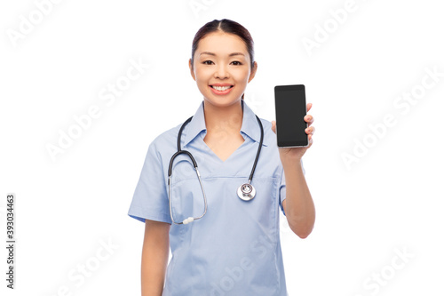 medicine, profession and healthcare concept - happy smiling asian female doctor or nurse in blue uniform with stethoscope showing smartphone over white background