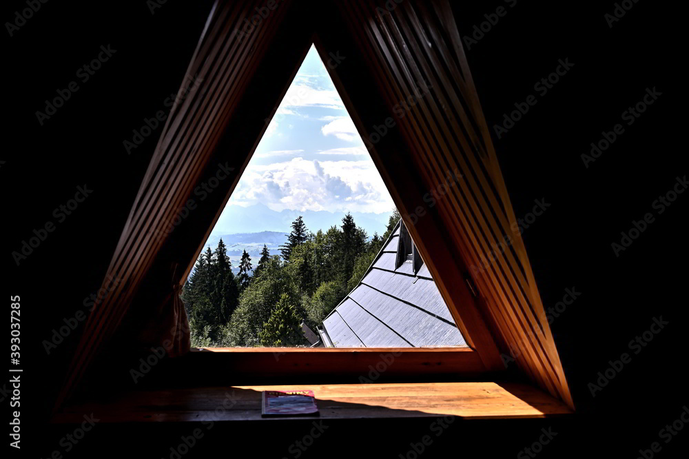 Beautiful view on green trees forest, blue sky and mountains through a wooden window frame of an old house cottage on a slope of a hill in southern Poland