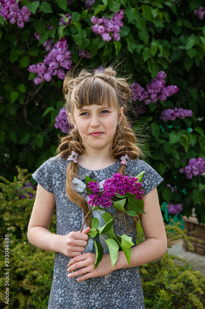A little girl with beautiful braids stands by a blooming tree, holding lilac flowers in her hands A little girl with beautiful braids stands by a blooming tree, holding lilac flowers in her hands