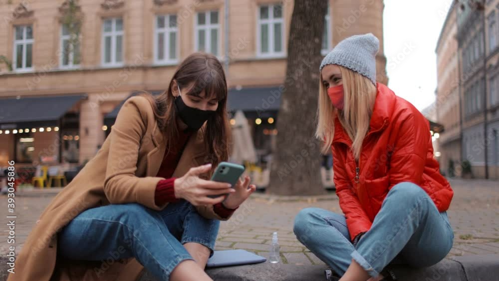 Joyful female friends smiling, chatting and using smartphone while sitting together outdoors. Young women wearing face medical mask during pandemic time. Joyful female friends smiling, chatting and using smartphone while sitting together outdoors. Young women wearing face medical mask during pandemic time.