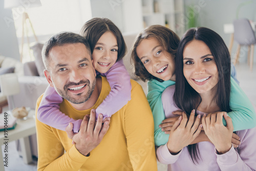 Photo of full family four people parents hold piggy back adopted kids shiny smile camera wear colorful sweater in living room indoors