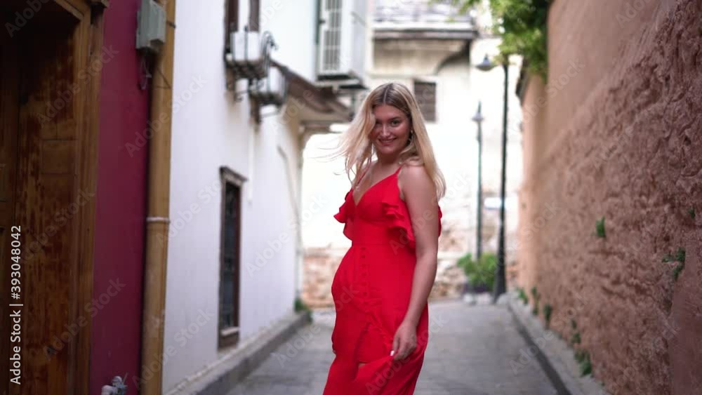 Nice young woman with charming smile and blonde straight hair in long red summer dress walks swirling along empty narrow street of ancient town slow motion. Nice young woman with charming smile and blonde straight hair in long red summer dress walks swirling along empty narrow street of ancient town slow motion.