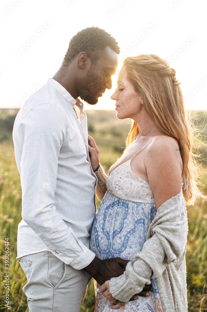 Tenderly multiracial pregnant couple enjoying time together outdoors, spouses in embrace in the rustic meadow Tenderly multiracial pregnant couple enjoying time together outdoors, spouses in embrace in the rustic meadow
