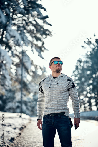 Young confident bearded man brutally posing in a winter forest in sunglasses and a knitted sweater