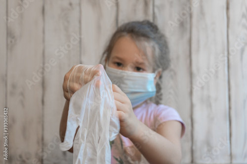 Little girl with a medical mask tries to put on disposable gloves spoiling the coronavirus, in a pink t-shirt, can't put on a glove, wooden photo background