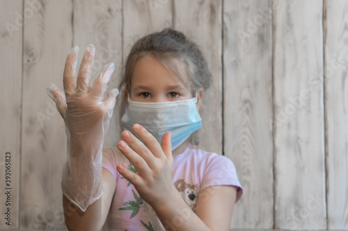 A little girl with a medical mask put on disposable gloves and admires how well she did, spoiling the coronavirus, in a pink t-shirt, can't put on a glove, wooden background photos
