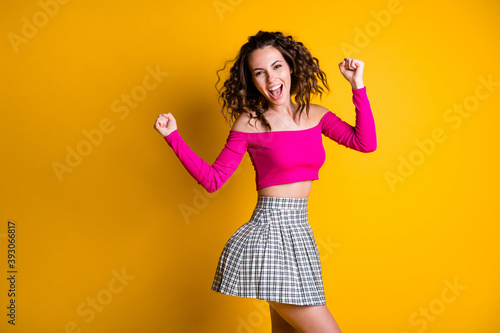 Photo portrait of excited young woman dancing holding fists up celebrating wearing fuchsia crop-top checkered skirt isolated on vivid yellow colored background