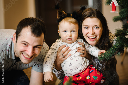 Mom, dad with child son play toys near the Christmas tree. Toddler boy in a Christmas costume and mother, father sitting on floor. Family winter holidays. Happy New Year and Merry Christmas.