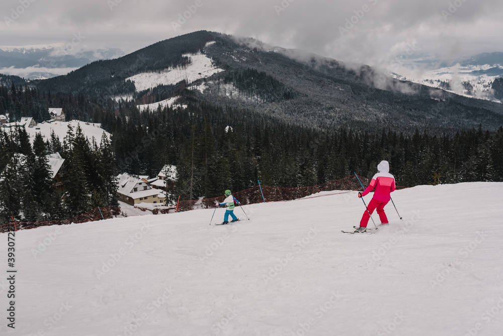 Woman and child on skis descend down from the mountain on snow track on background of forest. Skiing. Extreme winter sports. The skier goes downhill. Winter nature. Landscape. Top back view.