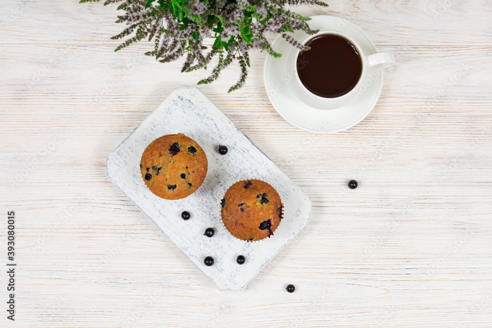 Cupcakes with black currants on a white wooden cutting board against the background of a cup of tea. View from above. Cupcakes with black currants on a white wooden cutting board against the background of a cup of tea. View from above.