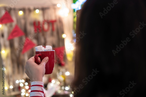 Woman hand with cup of hot cocoa with marshmallows at christmas time opposite small home cocoa bar.
