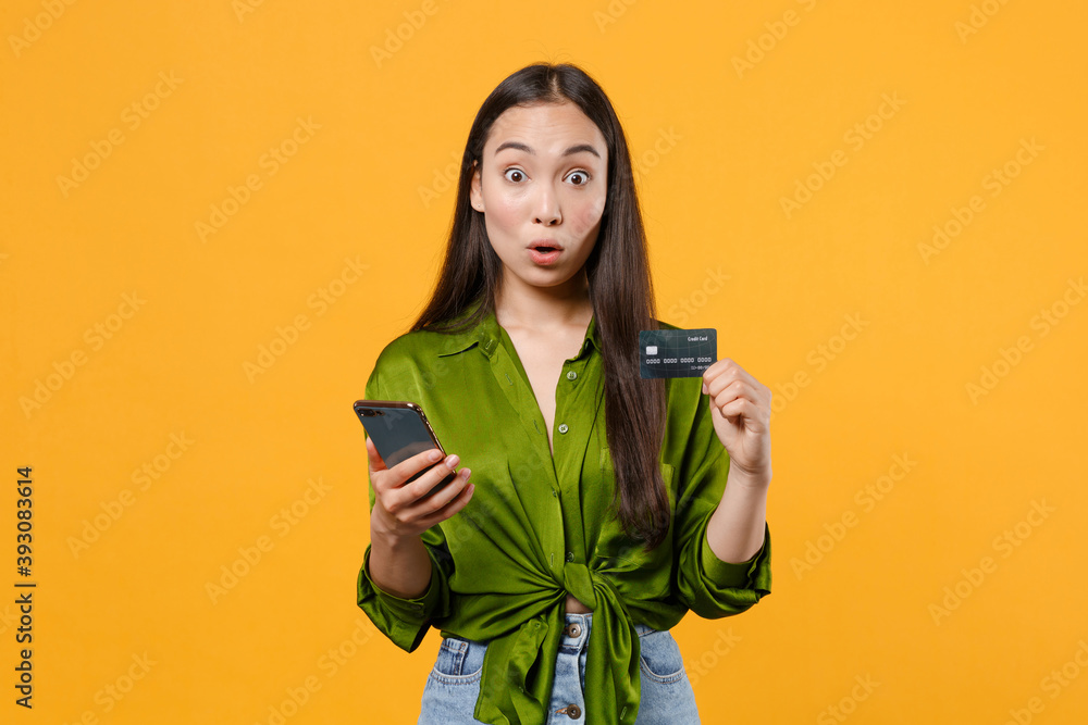 Shocked amazed young brunette asian woman wearing basic green shirt standing hold in hands using mobile cell phone hold credit bank card isolated on bright yellow colour background, studio portrait. Shocked amazed young brunette asian woman wearing basic green shirt standing hold in hands using mobile cell phone hold credit bank card isolated on bright yellow colour background, studio portrait.