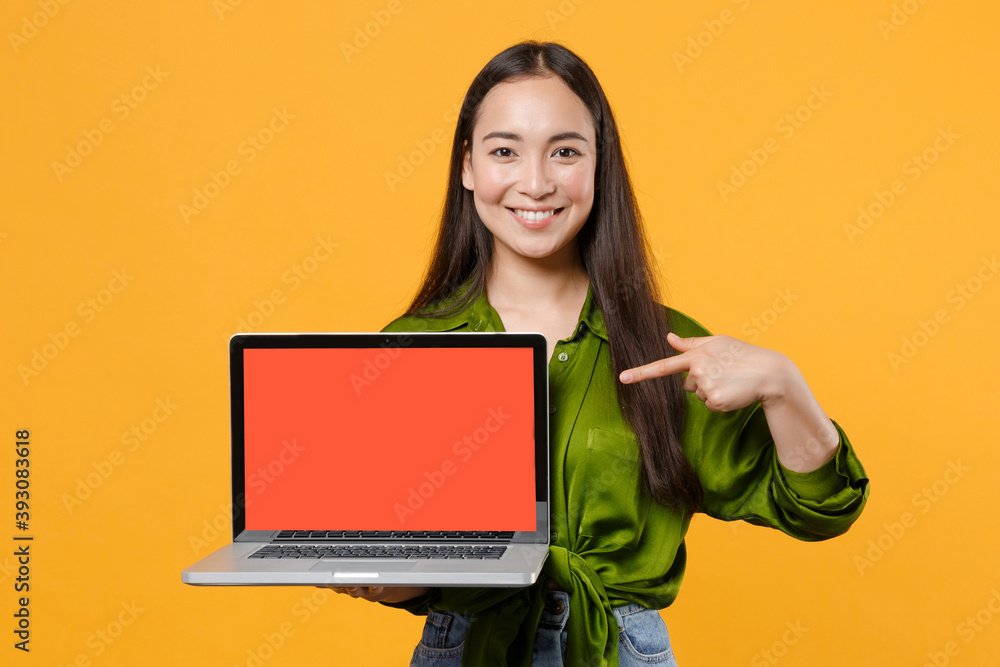 Naklejka premium Smiling young brunette asian woman in basic green shirt standing pointing index finger on laptop pc computer with blank empty screen mock up copy space isolated on yellow background, studio portrait.