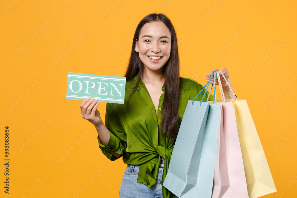Smiling young brunette asian woman 20s in basic green shirt standing hold package bags with purchases after shopping sign with OPEN title looking camera isolated on yellow background, studio portrait. Smiling young brunette asian woman 20s in basic green shirt standing hold package bags with purchases after shopping sign with OPEN title looking camera isolated on yellow background, studio portrait.