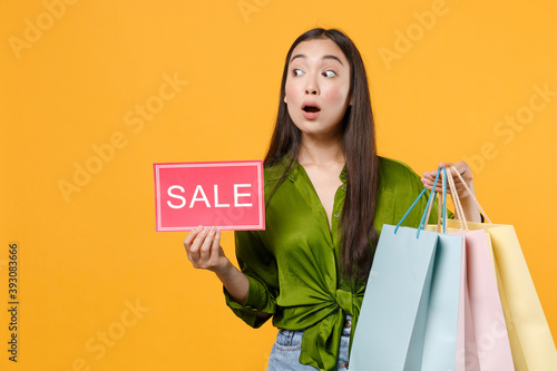 Shocked young brunette asian woman 20s in basic green shirt standing hold package bags with purchases after shopping sign with SALE title looking aside isolated on yellow background, studio portrait.