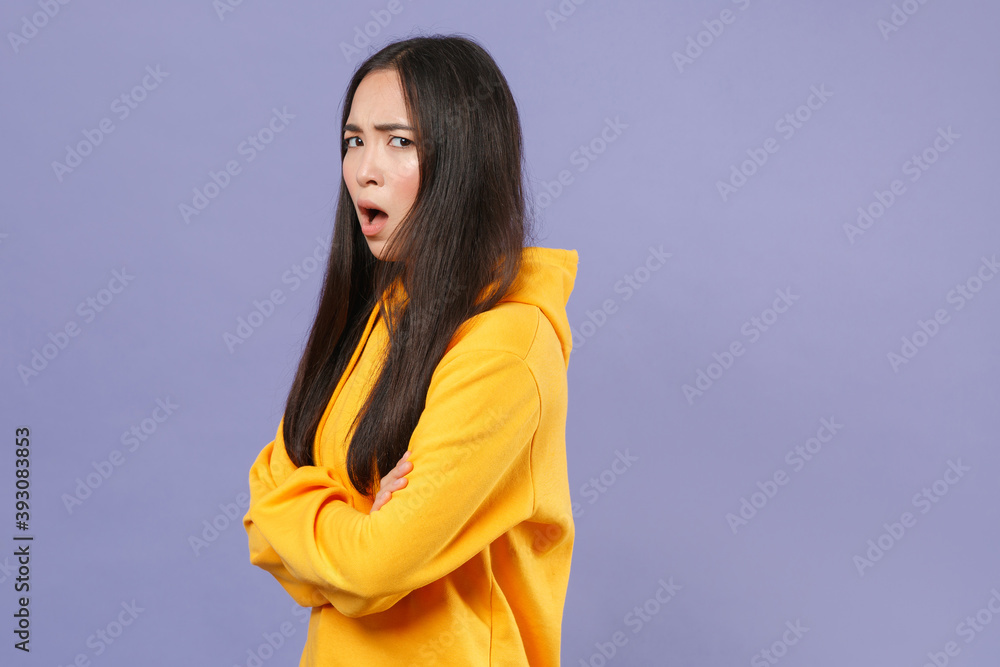 Side view of shocked worried young brunette asian woman wearing casual basic yellow hoodie standing holding hands crossed looking camera isolated on pastel violet colour background studio portrait. Side view of shocked worried young brunette asian woman wearing casual basic yellow hoodie standing holding hands crossed looking camera isolated on pastel violet colour background studio portrait.