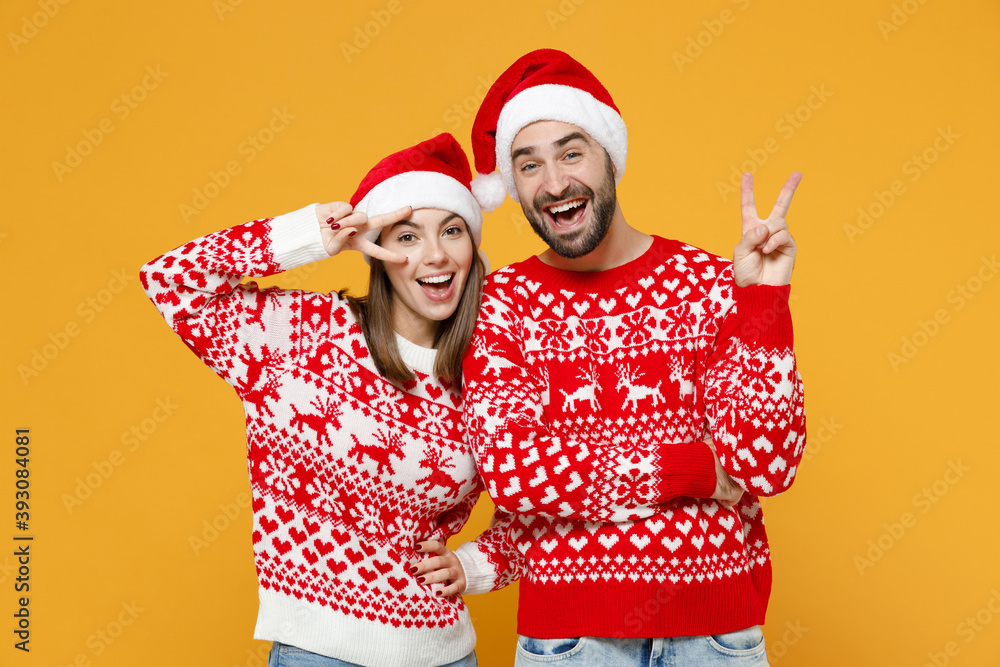 Funny young Santa couple friends man woman 20s wearing red sweater Christmas hat showing victory sign isolated on yellow background studio portrait. Happy New Year celebration merry holiday concept. Funny young Santa couple friends man woman 20s wearing red sweater Christmas hat showing victory sign isolated on yellow background studio portrait. Happy New Year celebration merry holiday concept.