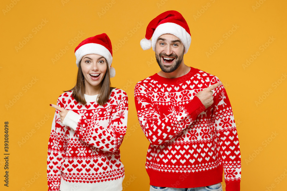 Excited young Santa couple friends man woman in red sweater Christmas hat pointing index fingers aside isolated on yellow background studio portrait. Happy New Year celebration merry holiday concept. Excited young Santa couple friends man woman in red sweater Christmas hat pointing index fingers aside isolated on yellow background studio portrait. Happy New Year celebration merry holiday concept.