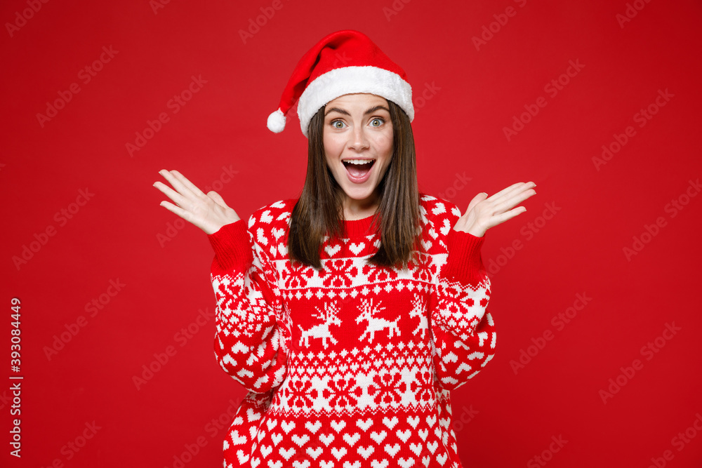 Surprised young brunette Santa woman in sweater, Christmas hat keeping mouth open spreading hands isolated on red colour background, studio portrait. Happy New Year celebration merry holiday concept. Surprised young brunette Santa woman in sweater, Christmas hat keeping mouth open spreading hands isolated on red colour background, studio portrait. Happy New Year celebration merry holiday concept.