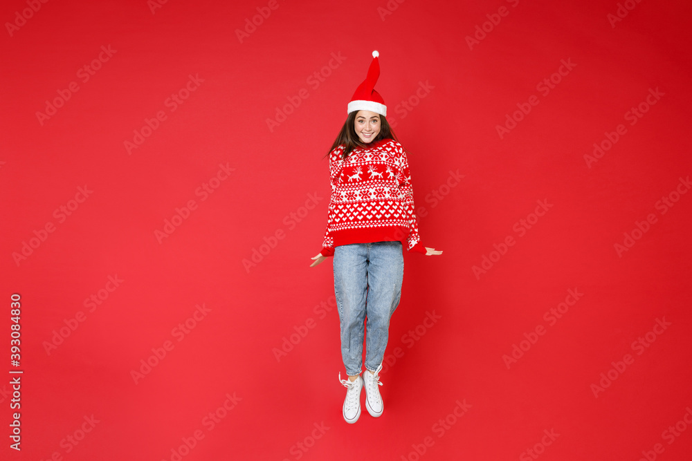 Full length of smiling young brunette Santa woman 20s wearing sweater, Christmas hat jumping having fun isolated on red background, studio portrait. Happy New Year celebration merry holiday concept. - obrazy, fototapety, plakaty 