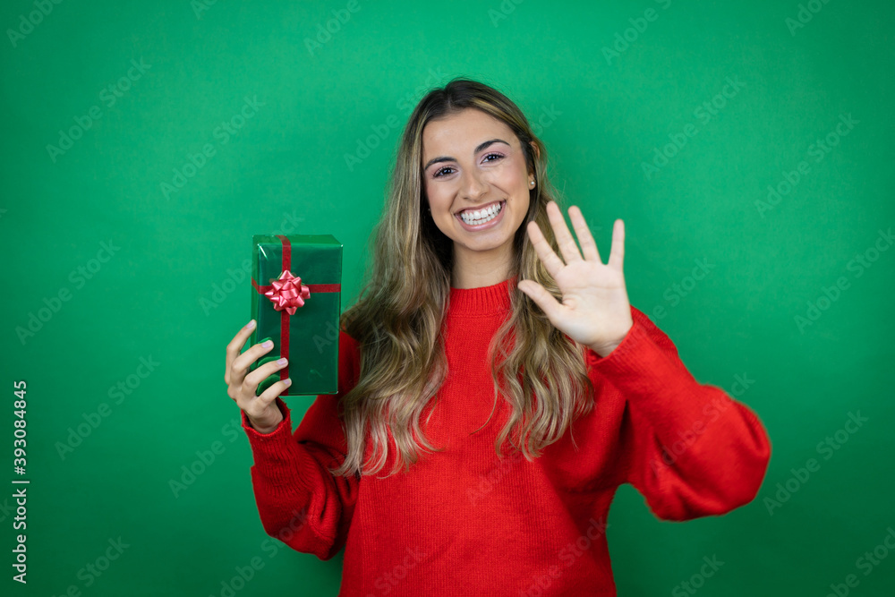 Young beautiful girl holding gift over isolated green background showing and pointing up with fingers number five while smiling confident and happy Young beautiful girl holding gift over isolated green background showing and pointing up with fingers number five while smiling confident and happy