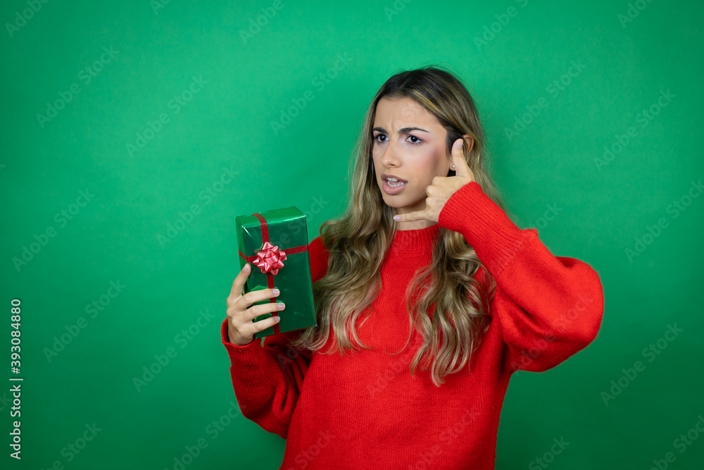 Young beautiful girl holding gift over isolated green background confused doing phone gesture with hand and fingers like talking on the telephone Young beautiful girl holding gift over isolated green background confused doing phone gesture with hand and fingers like talking on the telephone