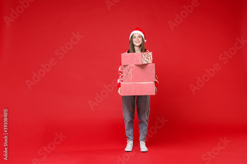 Full length of laughing young brunette Santa woman in sweater, Christmas hat hold present boxes with gifts isolated on red background studio portrait. Happy New Year celebration merry holiday concept. © ViDi Studio