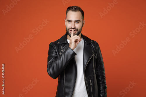 Secret young bearded man in basic white t-shirt black leather jacket standing saying hush be quiet with finger on lips shhh gesture looking camera isolated on orange colour background studio portrait.