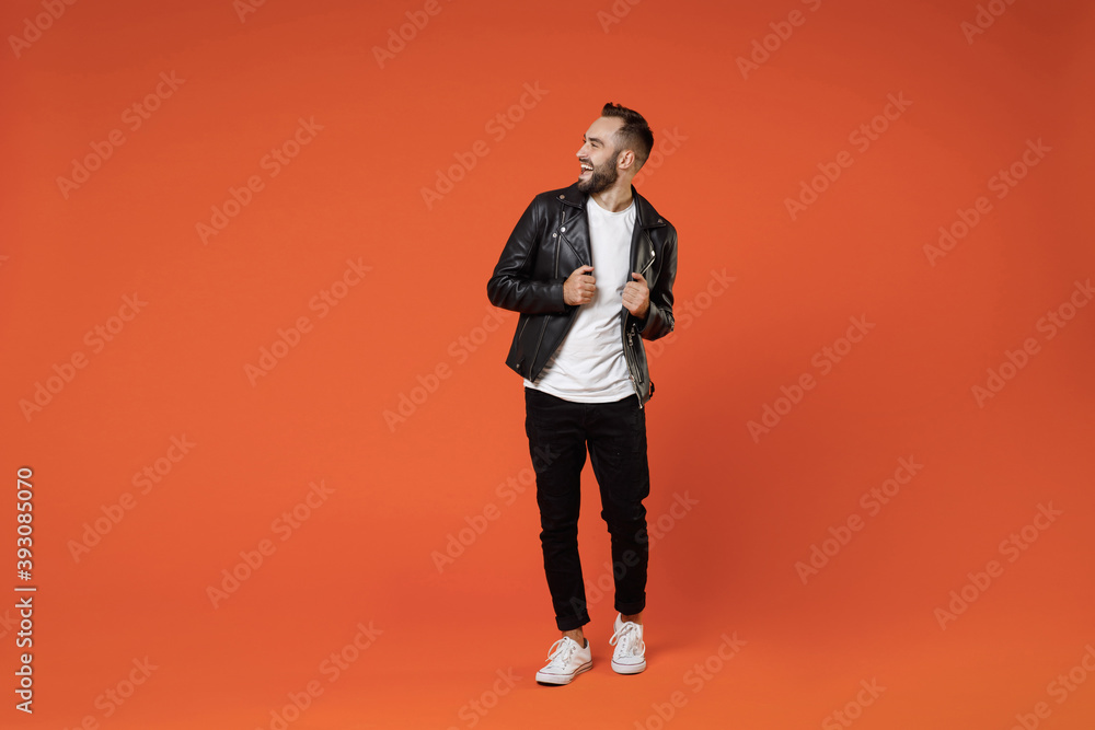 Full length of cheerful laughing funny excited young bearded man wearing basic white t-shirt black leather jacket standing looking aside isolated on bright orange colour background studio portrait. - obrazy, fototapety, plakaty 