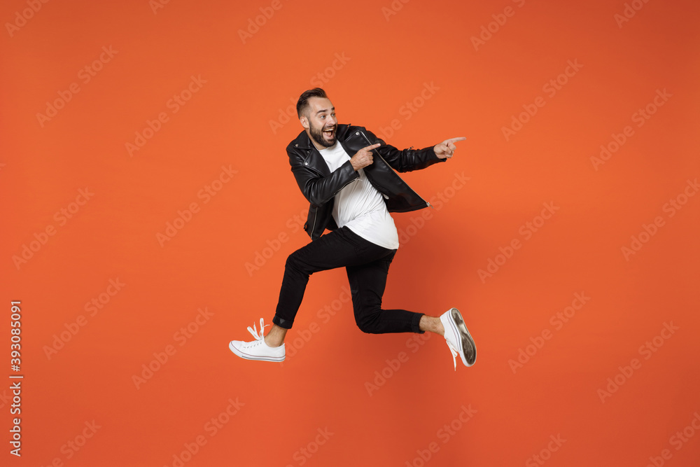Full length side view of excited surprised young bearded man 20s in basic white t-shirt black leather jacket jumping pointing index fingers aside isolated on orange colour background studio portrait.