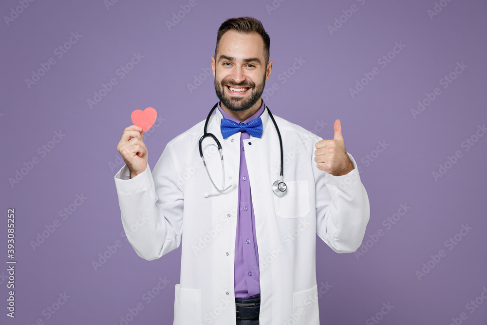 Smiling young bearded doctor man in white medical gown stethoscope hold red little heart showing thumb up isolated on violet background studio portrait. Healthcare personnel health medicine concept. Smiling young bearded doctor man in white medical gown stethoscope hold red little heart showing thumb up isolated on violet background studio portrait. Healthcare personnel health medicine concept.
