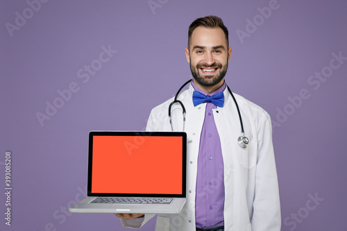 Smiling young bearded doctor man in medical gown stethoscope hold laptop computer with blank empty screen isolated on violet background studio portrait. Healthcare personnel health medicine concept.