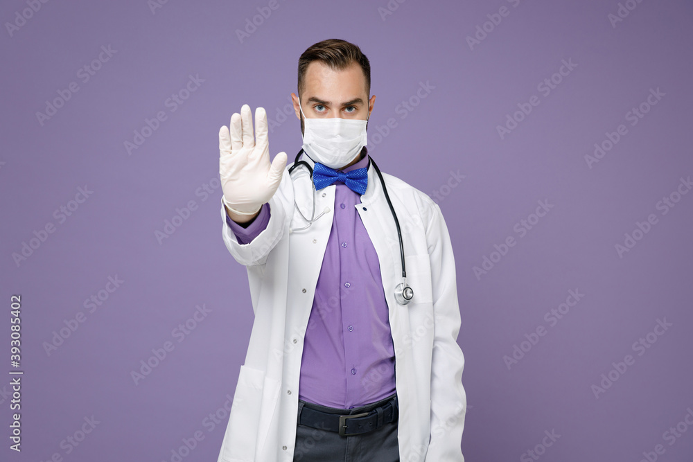 Young doctor man in medical gown sterile face mask to safe from coronavirus virus covid-19 showing stop gesture with palm isolated on violet background. Healthcare personnel health medicine concept. Young doctor man in medical gown sterile face mask to safe from coronavirus virus covid-19 showing stop gesture with palm isolated on violet background. Healthcare personnel health medicine concept.