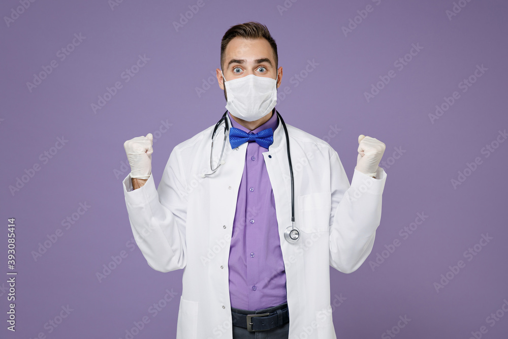 Amazed doctor man in medical gown stethoscope sterile face mask to safe from coronavirus virus covid-19 doing winner gesture isolated on violet background Healthcare personnel health medicine concept. Amazed doctor man in medical gown stethoscope sterile face mask to safe from coronavirus virus covid-19 doing winner gesture isolated on violet background Healthcare personnel health medicine concept.