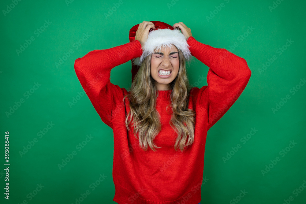 Young beautiful girl wearing Christmas Santa hat holding gift over isolated green background suffering from headache desperate and stressed because pain and migraine with her hands on head Young beautiful girl wearing Christmas Santa hat holding gift over isolated green background suffering from headache desperate and stressed because pain and migraine with her hands on head