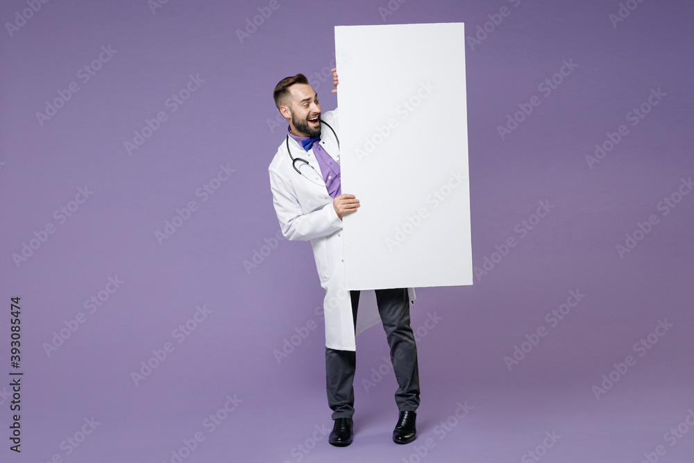 Full length of excited young bearded doctor man in white medical gown hold big white empty blank billboard isolated on violet background studio portrait. Healthcare personnel health medicine concept. Full length of excited young bearded doctor man in white medical gown hold big white empty blank billboard isolated on violet background studio portrait. Healthcare personnel health medicine concept.