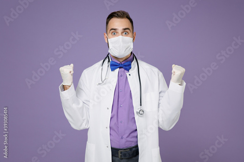 Amazed doctor man in medical gown stethoscope sterile face mask to safe from coronavirus virus covid-19 doing winner gesture isolated on violet background Healthcare personnel health medicine concept.