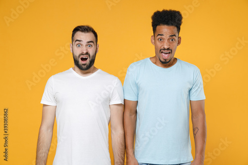 Shocked worried young two friends european african american men 20s wearing white blue casual t-shirts keeping mouth open looking camera isolated on bright yellow colour background studio portrait.